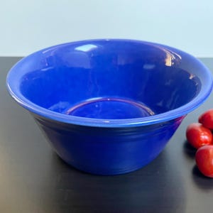 Peut inclure: Un saladier en céramique bleu cobalt avec un large bord. Le bol est vide et posé sur une surface sombre, avec un groupe de tomates cerises rouges à droite. La finition brillante du bol reflète la lumière.