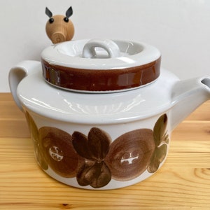 Könnte beinhalten: Weiße Keramik-Teekanne mit braunem Blumenmuster und passendem Deckel. Der Deckel hat ein braunes Band und einen weißen Griff. Die Teekanne hat einen Ausguss und einen Henkel. Eine Holzfigur eines Schweins befindet sich oben.