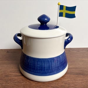 Puede incluir: Una olla de cerámica blanca y azul con tapa y asas. La olla tiene un patrón de hojas azules alrededor de la base y una pequeña bandera sueca en la parte superior. La olla está sobre una superficie de madera.