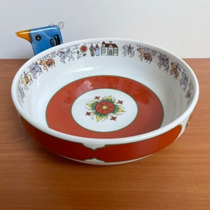 May include: White ceramic bowl with a rust-colored band and a floral design in the center. The rim features a repeating pattern of figures and buildings. A blue bird figurine is in the background.