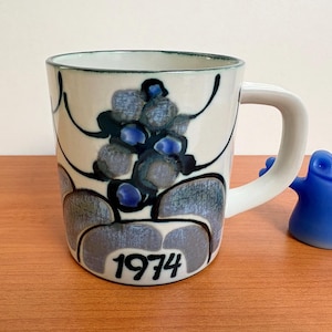 Può includere: Una tazza in ceramica con manico bianco e un motivo floreale nei toni del blu e del grigio. La tazza ha il numero "1974" stampato. Una piccola statuina blu è accanto alla tazza.