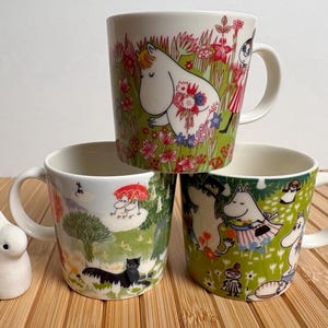 Op de afbeelding: Drie keramische mokken met kleurrijke Moomin-karakterillustraties en een klein wit keramisch konijnenbeeldje. De bovenste mok toont een wit wezen in een veld met bloemen, terwijl de onderste mokken verschillende scènes met Moomin-karakters laten zien.