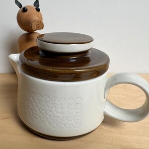 Op de afbeelding: Een witte keramische theepot met een bruin deksel en handvat. De theepot heeft een gestructureerd patroon op de voorkant en een gebogen handvat. Op de achtergrond staat een houten varkensbeeldje.