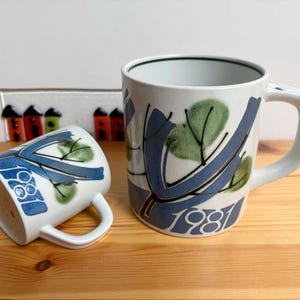 Può includere: Due tazze in ceramica bianca con disegni astratti blu e verdi e il numero 1981. Una tazza è in posizione verticale, l'altra è di lato. Le tazze hanno manici bianchi e sono su una superficie di legno.