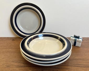 Könnte beinhalten: Stapel von vier cremefarbenen Keramikschalen mit marineblauem Rand und eine einzelne passende Schale. Eine kleine weiße Geschenkbox mit blauem Band ist ebenfalls vorhanden. Die Schalen stehen auf einer Holzoberfläche.