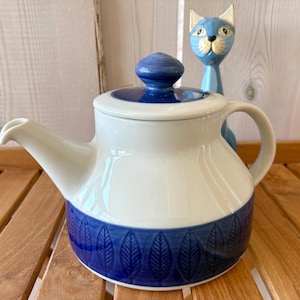 Könnte beinhalten: Weiß-blaue Keramik-Teekanne mit Deckel und Henkel. Die Teekanne hat ein blaues Band mit einem Blattmuster am Boden. Im Hintergrund befindet sich eine blaue Katzenfigur. Die Teekanne steht auf einer Holzoberfläche.