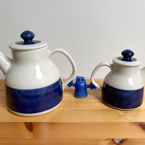 Peut inclure: Deux théières en céramique blanches et bleues avec un motif de feuilles, une grande et une petite, avec des couvercles assortis. Une petite figurine bleue se trouve entre les théières. Les théières sont sur une surface en bois.