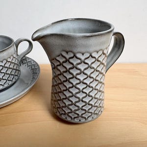 May include: A gray ceramic creamer with a handle and spout, alongside a matching teacup and saucer. The creamer and cup feature a repeating geometric pattern. The set is on a light wood surface.