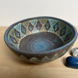 May include: A ceramic bowl with a textured interior and a diamond pattern in blue, tan, and white. The bowl has a dark brown exterior. Several polished stones are displayed beside the bowl. The bowl is likely handmade.