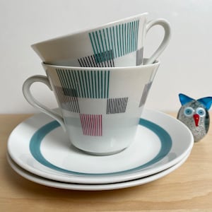 May include: Two white teacups with matching saucers. The cups and saucers have a teal and black geometric pattern. A teal stripe encircles the saucers. A small glass owl figurine is in the background.