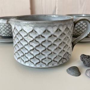Op de afbeelding: Een grijze keramische creamer met een handvat en tuit, met een textuur, ruitvormig ontwerp. Bijpassende kopjes en schotels zijn zichtbaar op de achtergrond. Decoratieve schelpen en een steen staan op de voorgrond. De set is voor thee of koffie.
