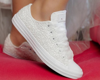 Könnte beinhalten: Weiße Converse-Sneaker aus Spitze mit einem weißen Satinband, das um den Knöchel gebunden ist. Die Sneaker sind mit weißen Perlen verziert und stehen auf einer roten Oberfläche.