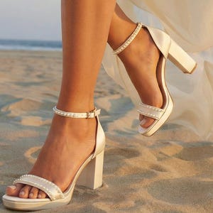 Könnte beinhalten: Weiße Satin-Blockabsatz-Sandalen mit Knöchelriemen, die mit kleinen, funkelnden Verzierungen verziert sind. Die offenen Schuhe haben einen dicken Absatz und werden an einem Sandstrand getragen. Ein fließender weißer Stoff ist sichtbar, was auf eine Hochzeit oder einen besonderen Anlass hindeutet.