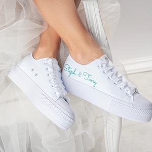 Pu&ograve; includere: Sneakers bianche in tela con lacci in raso bianco. La scarpa sinistra ha il testo "Steph & Tony" ricamato in turchese sul lato.