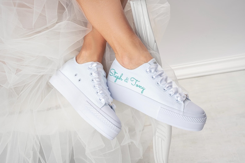 Pu&ograve; includere: Sneakers bianche in tela con lacci in raso bianco. La scarpa sinistra ha il testo "Steph & Tony" ricamato in turchese sul lato.