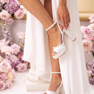 May include: A pair of white platform sandals featuring a thick block heel, an open toe design, and a delicate ankle strap adorned with small white beads. The shoes have a smooth finish and a secure buckle closure, providing a stable base for the foot.