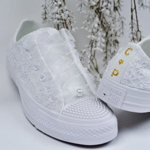 Peut inclure: Baskets Converse blanches recouvertes de dentelle avec des perles et des strass. Le bout est orné de petites perles et les lacets sont remplacés par un tissu transparent. Initiales dorées sur le talon.