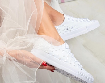 May include: White canvas sneakers embellished with pearls. The shoes have a classic low-top design with white laces and a rubber sole. The sneakers are decorated with small pearls and larger pearl accents.
