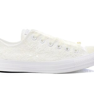May include: White lace Converse sneakers with a pearl accent on the side. The shoes have a white rubber sole and a white lace design.