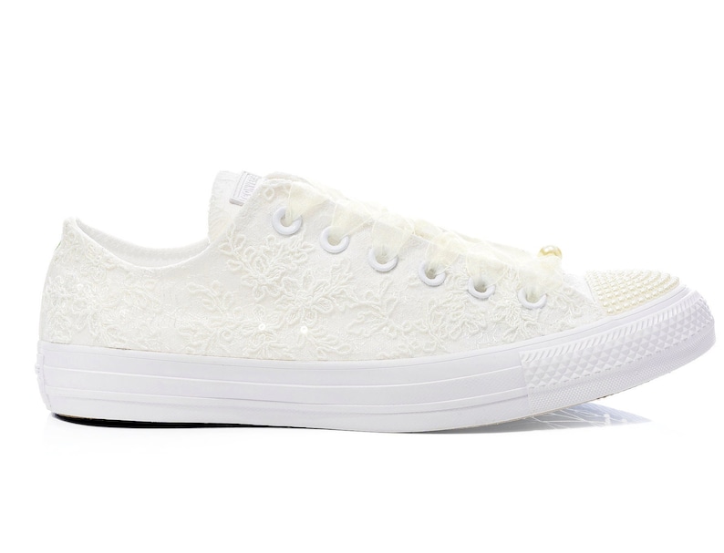 May include: White lace Converse sneakers with a pearl accent on the side. The shoes have a white rubber sole and a white lace design.
