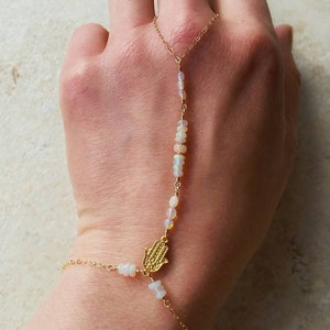 Könnte beinhalten: Eine zarte goldene Handkette mit Opalperlen und einem Hamsa-Hand-Anhänger. Die Kette verbindet einen Ring und ein Armband und schafft so ein einzigartiges Schmuckstück. Die Opalperlen sind klein und weiß und sorgen für einen dezenten Glanz.