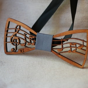 Peut inclure: Nœud papillon en bois avec un motif de notes de musique. Le nœud papillon marron clair présente des notes de musique et une clé de sol découpées. Il est maintenu par un ruban gris et une sangle noire.
