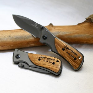 Peut inclure: Deux couteaux de poche avec des manches en bois. Les manches sont gravés avec le texte "Super témoin" et la date "04-04-2020".