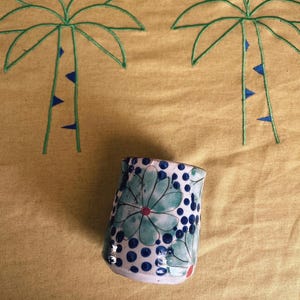 May include: A white ceramic mug with a blue and green floral pattern and red accents. The mug is sitting on a yellow fabric background with embroidered palm trees.