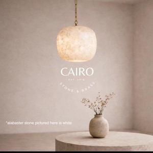 Pode incluir: Uma luminária pendente redonda de pedra de alabastro branco sujo pende de uma fina corrente de latão. Abaixo, um pequeno vaso de pedra contém hastes secas. Texto: CAIRO EST. 2016 STONE & BRASS. Nota: *alabaster stone pictured here is white.