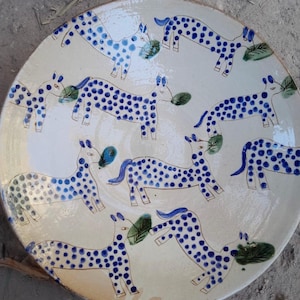 May include: A white ceramic plate with a blue and white design of horses. The horses are all facing the same direction and have blue spots on their bodies. The plate is round and has a slightly raised rim.