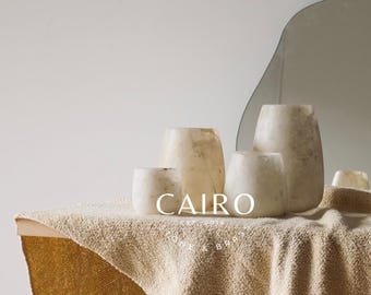 Puede incluir: Un conjunto de cuatro candelabros de alabastro blanco de diferentes tamaños, dispuestos sobre un mantel texturizado beige. El texto "CAIRO EST. 2016 STONE & BRASS" es visible en el centro de la imagen.