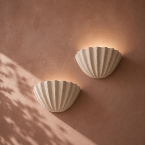 May include: Two off-white, semi-circular wall sconces with a textured, ribbed shell-like design. Each fixture is mounted to a wall and emits a soft, warm glow from the top, casting light upwards against the surface in a fan-shaped pattern.