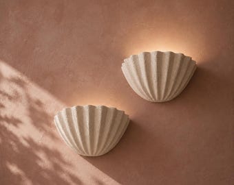 Può includere: Due applique semicircolari color bianco sporco con un design testurizzato a forma di conchiglia scanalata. Ogni lampada è montata a parete ed emette una luce calda e soffusa dalla parte superiore, proiettandola verso l'alto a ventaglio.