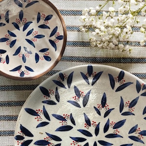 Könnte beinhalten: Zwei weiße Keramikteller mit einem blauen und roten Blumendesign. Die Teller liegen auf einer gestreiften Tischdecke.