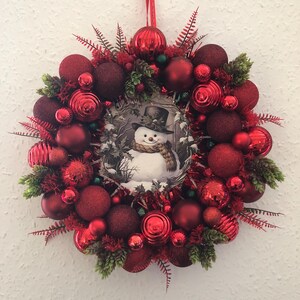 May include: A red Christmas wreath featuring a snowman illustration in the centre. The wreath is decorated with red baubles and green foliage.