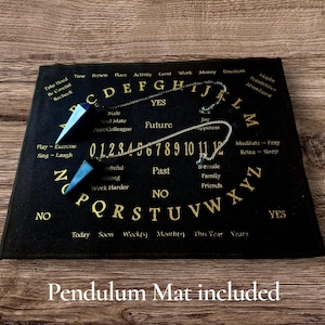 May include: Black Ouija board with gold lettering and a pendulum mat. The board features the alphabet, numbers, and words like "yes," "no," and "future." Two blue crystal pendulums are included. The text "Pendulum Mat included" is at the bottom.