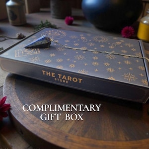 May include: A dark blue gift box with gold celestial designs and the text "THE TAROT STORE". A crystal pendulum hangs from the box. The words "COMPLIMENTARY GIFT BOX" are printed on the wooden surface below.