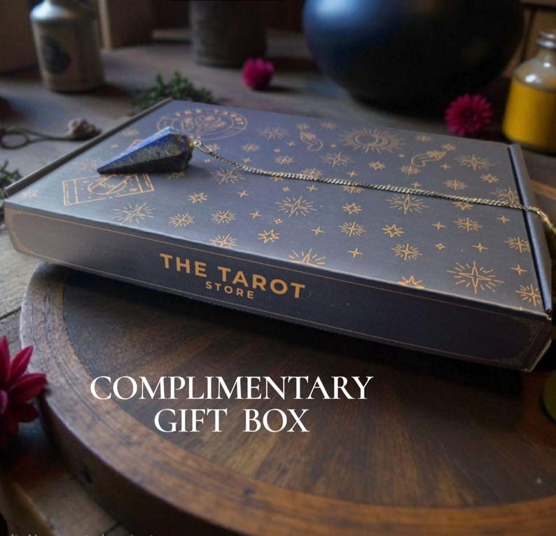 May include: A dark blue gift box with gold celestial designs and the text "THE TAROT STORE". A crystal pendulum hangs from the box. The words "COMPLIMENTARY GIFT BOX" are printed on the wooden surface below.