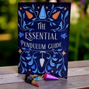 May include: A dark blue book titled "The Essential Pendulum Guide" with floral and geometric illustrations. Two pendulums, one striped and one pink, rest in front of the book. The book and pendulums are on a wooden surface.
