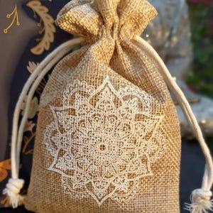 May include: A beige burlap drawstring bag with a white mandala design printed on the front. The bag has a natural, textured appearance and is tied with a white rope. The background is blurred, suggesting an outdoor setting.