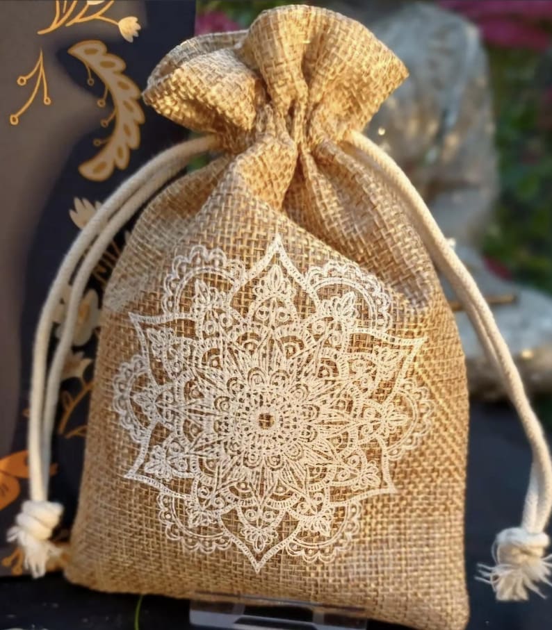 May include: A beige burlap drawstring bag with a white mandala design printed on the front. The bag has a natural, textured appearance and is tied with a white rope. The background is blurred, suggesting an outdoor setting.