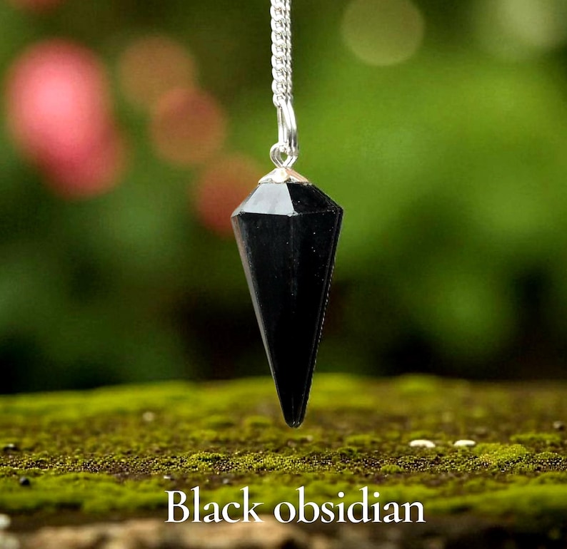 May include: A black obsidian pendant hangs from a silver chain. The pendant is a faceted, elongated cone shape. The text "Black obsidian" is at the bottom of the image. The background is blurred green and brown.