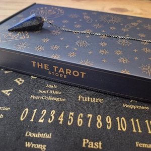 May include: A navy blue box with gold star and sun designs, and the text "THE TAROT STORE". A blue crystal pendulum hangs from the box. Below is a black cloth with gold numbers and letters.