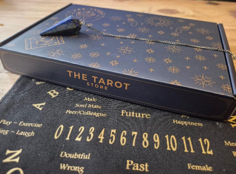 May include: A navy blue box with gold star and sun designs, and the text "THE TAROT STORE". A blue crystal pendulum hangs from the box. Below is a black cloth with gold numbers and letters.