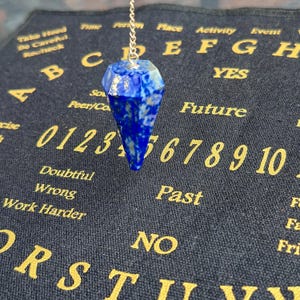 May include: A faceted, deep blue pendulum hangs from a silver chain. The pendulum is suspended above a dark blue Ouija board with gold lettering. The board features numbers, letters, and phrases like "Future" and "Yes."