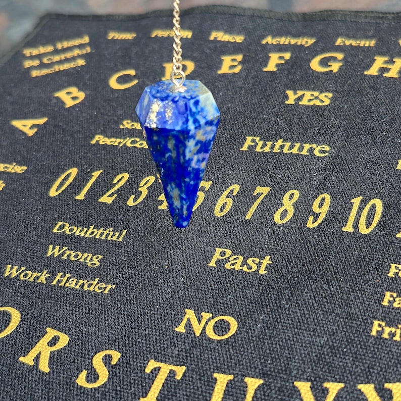 May include: A faceted, deep blue pendulum hangs from a silver chain. The pendulum is suspended above a dark blue Ouija board with gold lettering. The board features numbers, letters, and phrases like "Future" and "Yes."