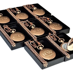 Puede incluir: Ocho relojes de bolsillo de oro con cadenas en cajas negras. Cada reloj tiene un monograma grabado diferente en la parte delantera. Los relojes est&aacute;n dispuestos en un patr&oacute;n escalonado.