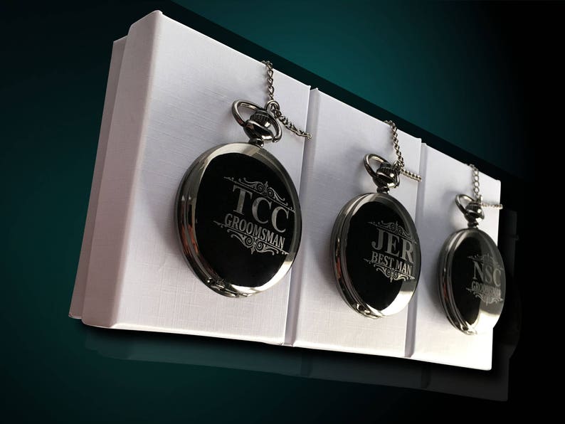 Puede incluir: Tres relojes de bolsillo plateados con caras negras y texto grabado. Los relojes se muestran en cajas blancas con un fondo verde oscuro.