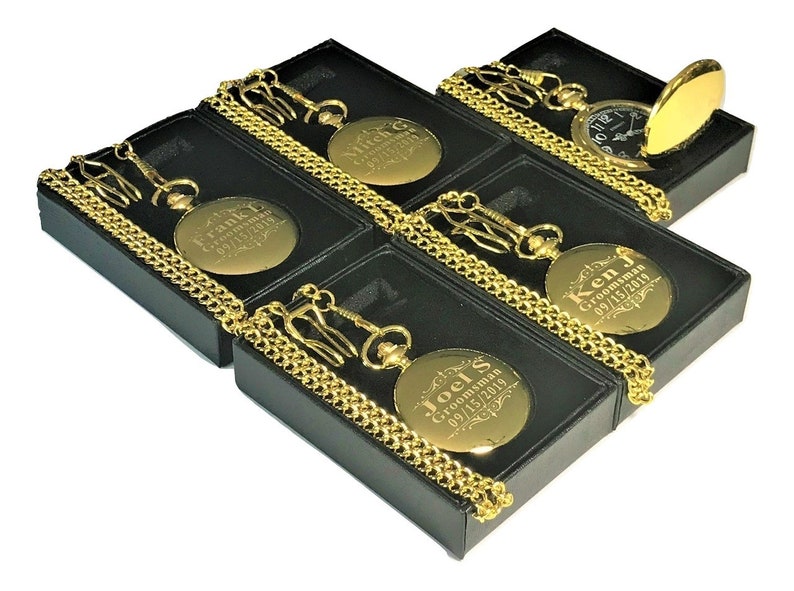 Puede incluir: Cinco relojes de bolsillo dorados con cadenas en cajas negras. Cada reloj tiene una inscripci&oacute;n personalizada con el t&iacute;tulo "Groomsman" y una fecha.