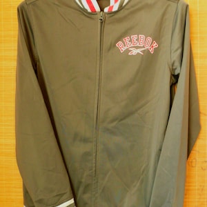 May include: An olive green zip-up track jacket featuring a full-length front zipper, long sleeves, and a ribbed collar with red and white stripes. The word REEBOK is embroidered in red on the chest, and the cuffs and hem have red and white stripes.